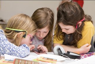 3 female children looking intently at a smartphone. 2 are wearing hearing devices and there is a pair of headphones on the table.