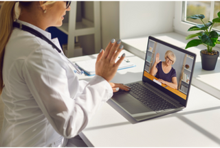 A doctor wearing a white coat waving at a patient on a video call using a laptop